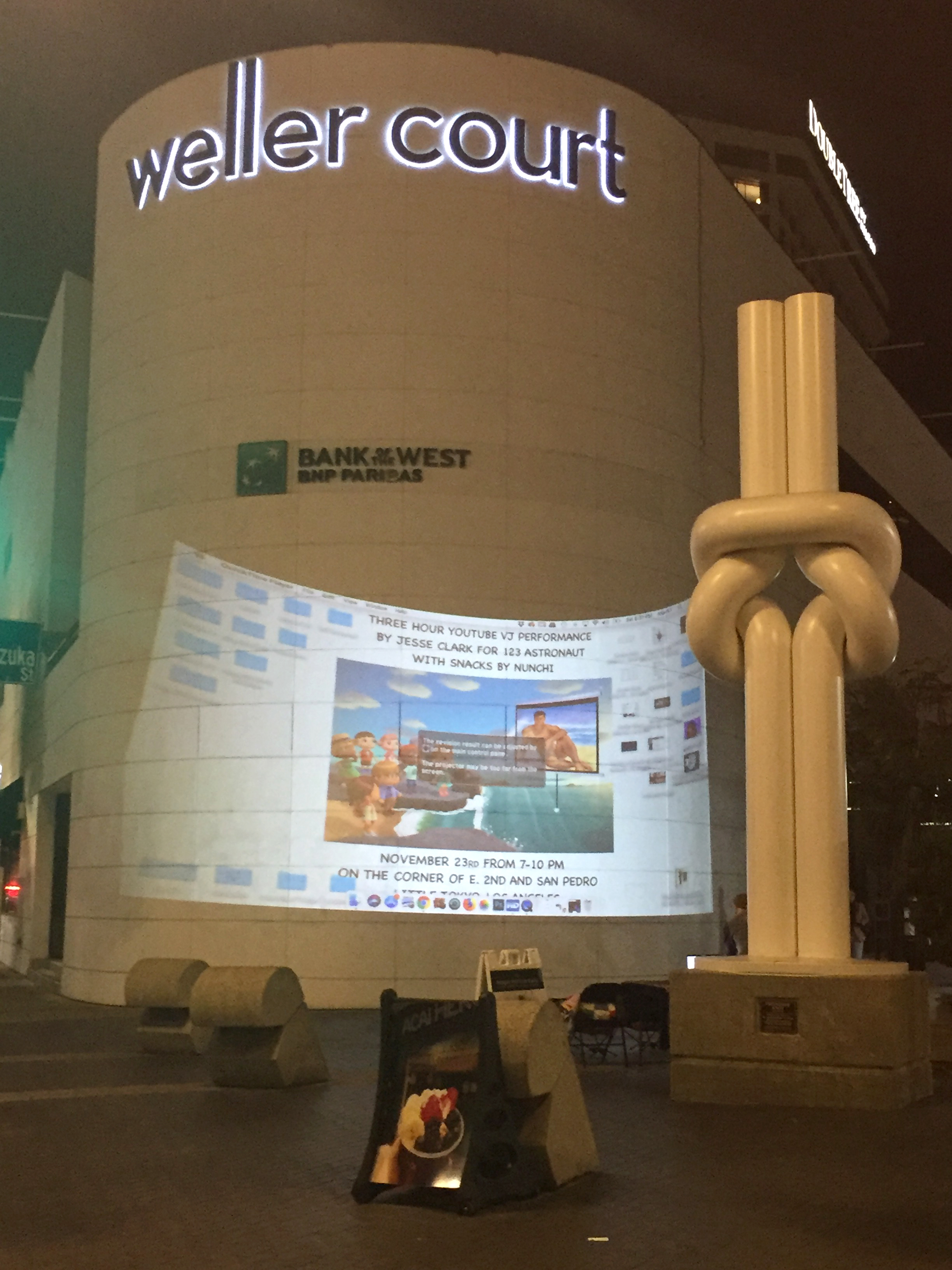 A corner in Little Tokyo, Los Angeles with a projection on a bank building next to a sculpture of a white knot